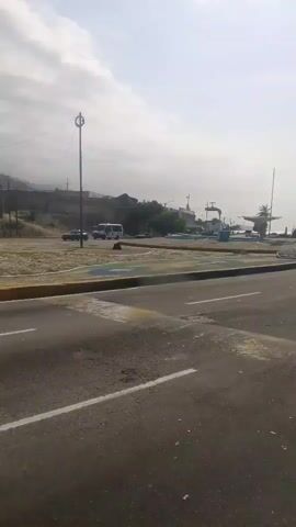 The camera is looking toward a paved roadway, which is divided by double white lines. There is a median to the left of the road with a short concrete barrier around it. The median is decorated with colorful paint and appears to be newly constructed.  In the distance, buildings, vehicles, and palm trees are visible against a light gray sky. Music is playing in the background. The video appears to have been taken from inside a moving vehicle.