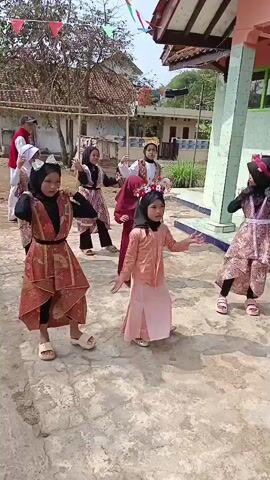 A group of young girls, most wearing headscarves and traditional attire, are performing a dance outdoors. The ground is paved with cobblestones, and a simple building with a red roof is visible in the background, along with trees and a blue tarp. Some of the girls are wearing floral headbands. One girl in the front center is wearing a peach-colored outfit and has a flower crown. Another girl to her right is wearing a pink and brown patterned dress and has pink cat ears. The girls are arranged in a semi-circle, facing towards the camera. The weather appears to be sunny. A man in a red vest and white shirt is standing in the background, appearing to observe the dance. Festive flags are strung across the top of the frame.