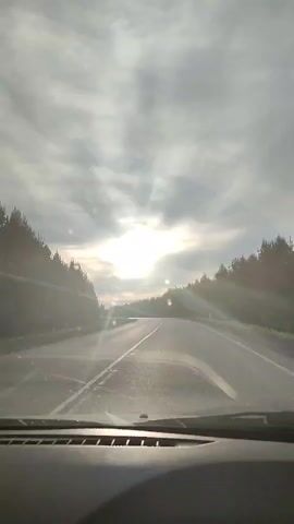 A video shows a first-person perspective of a car driving down a highway. The sky is cloudy, with the sun shining brightly through the clouds. Trees line the sides of the road. The car's dashboard is visible at the bottom of the frame. The sound of music is audible.