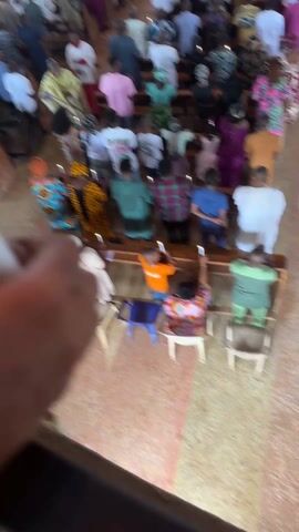 The video depicts a church service, likely Catholic, seen from an elevated angle. The focus is on a hand holding a lit white candle. Below, the congregation stands in pews, many holding lit candles. A priest in a gold robe is seen at the front. People are dressed in diverse clothing, including traditional African prints. The church interior features wooden pews and a tiled floor. The atmosphere appears somber and respectful.