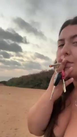The video starts with a woman holding a lit cigarette and keys, standing on a sandy area near a grassy dune, with the ocean visible in the background under a cloudy sky. The camera then pans to the right, showing a wider view of the sandy area leading to the ocean, with a fence running along the side. The sky is mostly overcast.