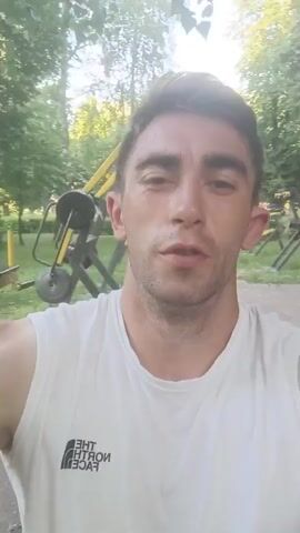 This is a selfie-style video of a young man in Fastiv, Ukraine. He appears to be outdoors, possibly in a park or recreational area, with trees and greenery visible in the background. The lighting suggests it is daytime, likely late afternoon or early evening due to the warm tones.

The man is wearing a white, sleeveless t-shirt with the "The North Face" logo visible on the left chest area. He is focused on the camera, and his expression suggests he is talking or reacting to something. In one segment, he flexes his bicep, indicating some sort of physical activity or display of strength. The overall mood seems casual and possibly energetic, given the implied outdoor setting and the flexing action. There are no other people or specific objects prominently featured besides the man and his clothing. The environment is somewhat blurred, characteristic of a video taken while moving or with a shallow depth of field.