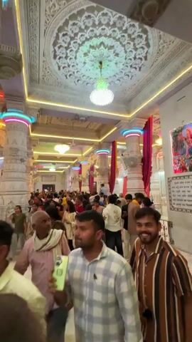 The video shows a crowded interior space. The space is adorned with white, ornately carved columns and ceilings. Many people are walking through the space, with some holding up phones to take pictures or videos. The people appear to be a mix of ages and are dressed in various clothing styles. There are colorful lights highlighting the architecture. The general movement suggests a religious or cultural site with a large number of visitors.
