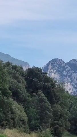 The video pans across a scenic mountainous landscape under a clear blue sky. The foreground is dominated by lush green trees, with a winding road visible on the left. A utility pole stands prominently on the right side of the frame. In the background, rugged, gray, and blue-hued mountains stretch across the horizon, their rocky textures clearly defined. The terrain suggests a rural or natural setting, possibly a hiking or driving route through a mountainous region. There is no visible text in the video. The lighting indicates it is daytime, likely morning or afternoon, with good visibility. The overall impression is one of natural beauty and tranquility.