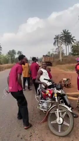 The video opens to show a group of people standing by the side of a road. Most of the men in the group are wearing maroon shirts. One man is sitting on a red motorcycle, posing and smiling. A few women in colorful dresses and headscarves are standing nearby. The camera pans to show a wider view of the scene, including a dirt road and some buildings. In the background, there's a truck and some palm trees. The overall environment appears to be a rural area.