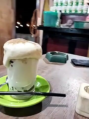 A tall glass of white liquid, topped with a white foamy substance and a light brown powder, sits on a green saucer on a wooden table. A black straw and a small silver spoon are also on the saucer. A mobile phone is on the table to the right of the glass. Behind the table is a counter, a wooden chair and shelves with water bottles.