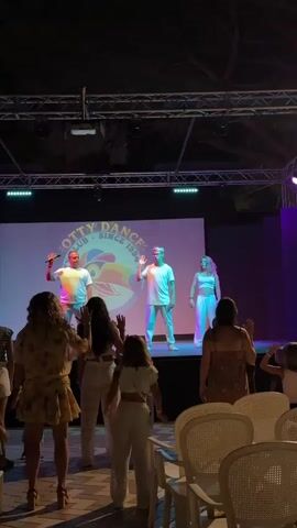 A group of four performers are on a stage, illuminated by purple and white stage lights. They are all wearing casual white clothing and appear to be dancing or leading an audience in a dance. The backdrop behind them is a large screen displaying the logo "OTTY DANCE CLUB SINCE 1994" with a colorful graphic. In front of the stage, a group of audience members, mostly women and girls, are standing and facing the stage with their hands raised, as if participating in the dance. The scene appears to be outdoors at night, with trees visible in the background and some tables and chairs in the foreground.