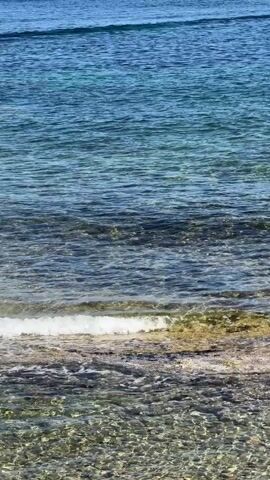 The video shows a rocky shoreline meeting the ocean. Small waves lap at the shore, and the water is clear and turquoise in color. The sky is blue. There are no people or other objects visible in the scene. The focus is solely on the natural environment.