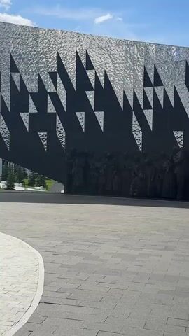 The video pans across a wall sculpture depicting a crowd of people, likely a memorial. The figures are rendered in dark, monochromatic relief against a textured, metallic wall with a geometric, jagged pattern. The people appear to be of various ages and genders, some holding children, others with bowed heads, suggesting themes of grief or remembrance. The ground in front of the sculpture is paved with grey rectangular tiles. The sky above is blue with scattered white clouds. The lighting suggests an outdoor setting on a bright day.