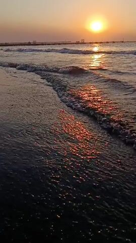A video shows a close-up view of gentle ocean waves rolling onto a dark, wet beach at sunset. The sun is a bright orb in the sky, casting a shimmering golden reflection across the water. In the distance, a long pier extends out into the sea with a few structures visible on it. The sky is clear and a soft orange-pink hue, suggesting the sun is low on the horizon. The camera angle is low, emphasizing the movement of the water and the texture of the wet sand. There are no people or animals visible in the frame. The scene is peaceful and serene.