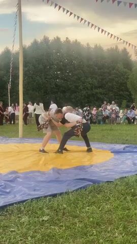 Two men are wrestling on a yellow mat surrounded by a blue tarp outdoors. The men are wearing casual clothing, and the taller man is lifting the shorter man up. There is a crowd of people watching the match, and a pole with flags is visible in the background. The event appears to be a festival or fair, with trees and a cloudy sky in the distance.