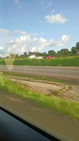 The video shows a roadside view from inside a moving vehicle on a sunny day with scattered clouds. The road is paved, with a grassy median and a dirt shoulder. The vegetation is lush and green. Three cars are parked on the far side of the road. A white car passes in the opposite direction, followed by a white truck. A billboard is visible in the background. The vehicle filming is moving quickly, as evidenced by the blurry foreground. Music is playing.