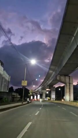 A time-lapse video shows a road under an elevated structure, possibly a metro line, as dusk settles. The sky transitions from a light blue with white clouds to a deep purple with darker clouds. Streetlights and lights from passing vehicles illuminate the road, which has white lane markings. A few people are visible walking and riding motorcycles on the road. The elevated structure is made of concrete and supported by pillars. Signs are visible on the side of the road and on the pillars. The scene is captured from a moving vehicle.