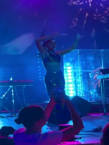A woman in a sparkling green two-piece outfit is performing on a stage under purple and blue lighting. She is dancing and moving her arms in a choreographed manner. The stage is equipped with musical instruments, including keyboards and drums, and there are lights illuminating the background with abstract patterns. In the foreground, a person is holding up a smartphone to record the performance.