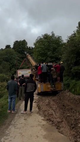 A group of men are gathered around a yellow crane truck on a muddy dirt road. The crane is lifting a white trailer with "Butola TATA Blow Hor" written on it. The men appear to be working together to manage the trailer and the crane. Some men are standing on the back of the truck, while others are on the ground. The environment is a rural, hilly area with trees and foliage. The sky is overcast.