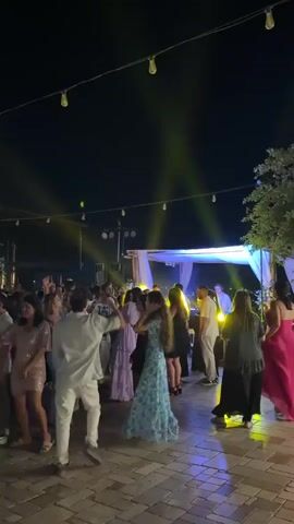 A nighttime outdoor party is in progress. A crowd of people are dancing on a paved area. String lights hang overhead, and stage lights illuminate the scene. Some people are wearing formal attire. The atmosphere is lively and festive.