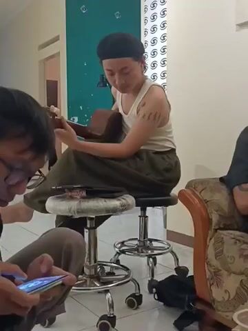 Here is a concise description of the video: 

The video captures an indoor scene with three individuals. In the center, a person sits on a wheeled stool, playing a brown guitar. This individual wears a white tank top, an olive-green skirt, and a black head covering. A tattoo is visible on their left arm. To their left, another person is seated on the floor, engrossed in their phone. On the right side of the frame, a third person is partially visible, seated on an upholstered chair.

The room has cream-colored walls. A partially visible green wall is behind the person playing the guitar. A door is also seen on the left of the frame.