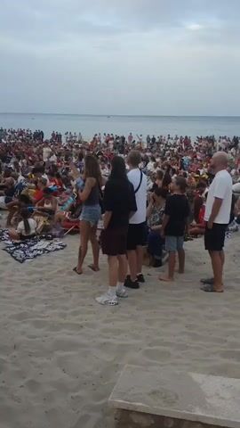The video shows a large crowd of people gathered on a sandy beach, facing the ocean. The sky is overcast, and the water is calm. Most of the people are sitting on blankets or chairs, and some are standing. Many people are wearing casual clothing, and some are holding Spanish flags. There are also a few palm trees visible in the background. The overall impression is of a public gathering or event, possibly a concert or festival. The camera is handheld and moves slightly throughout the recording, suggesting it was filmed by someone in the crowd.