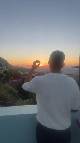 In a captivating 6-second video, a person is seen from behind, standing on an elevated viewpoint, gazing at a breathtaking sunset over a city or town. The sky is a gradient of clear blue transitioning to warm orange and red hues as the sun dips below the horizon. The setting is an outdoor landscape, likely a mountainous or hilly region, with lush green foliage and some buildings visible in the midground, contrasting with the expansive urban sprawl in the distance.

The individual, a man wearing a white short-sleeved shirt, initially holds his hand up, framing the setting sun between his thumb and forefinger as if "holding" it. He then lowers his hand and turns his head slightly to the right, revealing a side profile and a warm smile, seemingly enjoying the beautiful view. The scene evokes a sense of peace and wonder. Given the provided context, the location is Tétouan, Morocco. No visible text is present in the video.