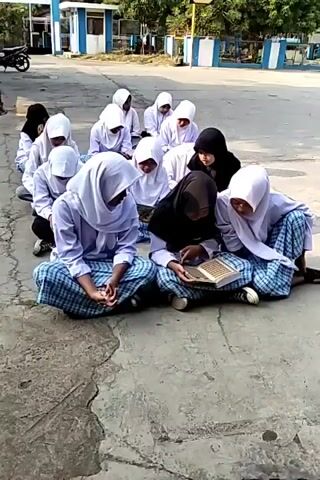 A group of young people, likely students, are sitting on the ground in rows. They are all wearing white headscarves and light-colored long-sleeved shirts, and checkered skirts or pants. In the front row, two individuals are leaning forward, one of whom is holding and looking at a book. The background appears to be an outdoor area with a paved surface and some buildings with blue and white accents. The lighting suggests it is daytime.