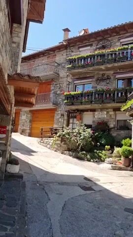 This is a video of a charming, historic stone building in a European village. The building has multiple stories with traditional architecture, including wooden balconies adorned with planters filled with red flowers. Lush green vines climb up the stone facade, adding to the picturesque scene. The street in front of the building is made of cobblestones, indicating an older European setting.

There are no people visible in the video, but there are several potted plants and some outdoor seating, suggesting a welcoming atmosphere, perhaps a small guesthouse or cafe. The time of day appears to be sunny, likely late morning or early afternoon, given the bright light and clear blue sky. The weather seems pleasant.

A sign visible on the building reads "CAN ROTA APARTAMENTS". The overall impression is one of tranquility and rustic beauty, characteristic of a quaint European village. The architecture and natural elements create a visually appealing and serene environment.