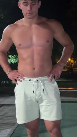 A shirtless person stands with hands on their hips, wearing light-colored shorts. The person then raises their arms above their head and stretches. The scene appears to be outdoors at night, with some foliage and lights visible in the background.