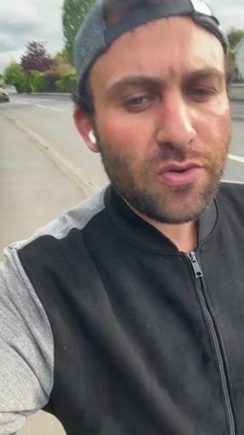 A man with a beard and a gray baseball cap walks along a street, filming himself with a handheld device. He is wearing a black jacket with gray sleeves. The street has sidewalks and is lined with houses and hedges. The sky is overcast.