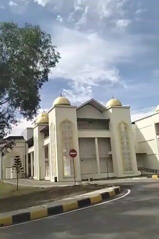 The video shows a slow-moving shot of a light-colored building with gold domes, possibly a mosque, seen from a roadside. There are several trees in the foreground and midground, partially obscuring the view of the building. The sky is partly cloudy. The road in the foreground has yellow and black striped curb markings. A red and white circular sign with a horizontal bar is visible on the side of the building. The camera is moving to the left, from the perspective of someone traveling forward.