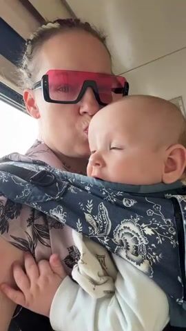 A woman is holding a baby in a carrier. The woman is wearing large pink sunglasses and a pink floral shirt. She has her hair in braids. The baby is looking to the side with blue eyes. They are in what appears to be a train or bus. The scene is brightly lit, suggesting daytime. The woman is puckering her lips as if to kiss the baby. The overall mood seems affectionate and happy. There is no visible text in the frame. The location context is Grushevskaya, Russia.