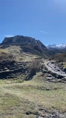 The video displays a rugged mountainous landscape with a clear blue sky overhead. In the foreground, the terrain is uneven and covered with patches of green grass, dry vegetation, and rocky areas. A winding dirt path meanders through the landscape. In the mid-ground, a rocky cliff formation is visible on the left side of the frame, while the background showcases snow-capped mountain peaks against the horizon. The scene is bathed in bright sunlight, creating a sharp contrast between the light and shadow on the landscape.