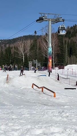 The video depicts a ski resort during the daytime under a clear blue sky. The foreground is a snow-covered area with several orange and black rails designed for snowboarders and skiers to perform tricks. Several individuals are engaged in snowboarding and skiing activities, attempting to slide along the rails. A chairlift with enclosed cabins is visible in the background, along with a slope dotted with skiers and snowboarders. Evergreen trees line the edge of the slope. In the mid-ground, there is a colorfully painted support structure for the chairlift, showing a graphic image of a sun.  The overall scene shows the lively activity of a winter sports resort.