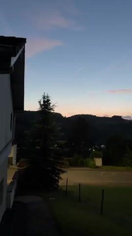 A video shows a twilight scene with a clear sky. In the foreground, a dark silhouette of a building and a tall evergreen tree are visible. A grassy lawn with a small decorative tree adorned with lights is in the middle ground. A street runs horizontally across the frame, with parked cars and a few houses visible on the other side. In the background, dark, tree-covered hills rise, with a castle-like structure illuminated on the hilltop. The sky displays subtle pink and orange hues near the horizon, transitioning to a deep blue overhead, with faint contrails from aircraft visible.