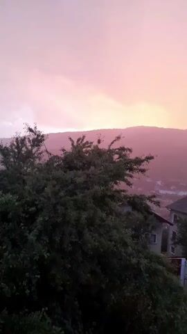 A video shot from a window overlooks a tree-filled hillside with houses nestled below. The sky is a soft pink and orange, suggesting either sunrise or sunset. A mountain range is visible in the distance.