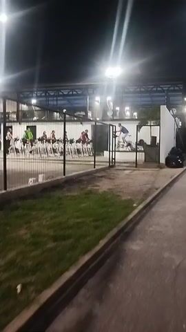 The video depicts an outdoor gym at night, illuminated by overhead lights. A row of stationary bikes are occupied by individuals engaged in cycling, with one person standing on their bike. A chain-link fence separates the gym area from a concrete path and a small patch of grass. The camera appears to be held and moves slightly. The overall atmosphere suggests an active fitness session in a well-lit outdoor environment.