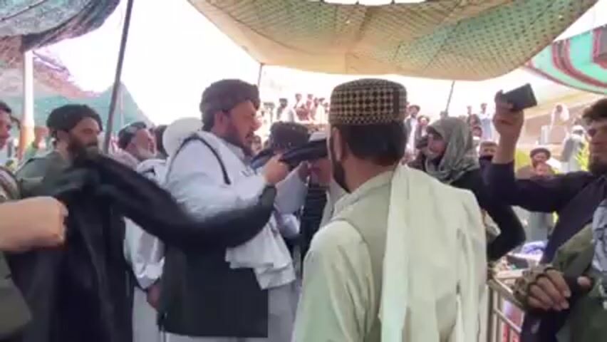 Here is a detailed description of the video:

**Scene:** The video appears to be taken at an outdoor event, possibly a gathering or ceremony, under a makeshift tent or canopy providing shade.

**People:**
*   A large group of men are present, many wearing traditional Afghan clothing, including turbans and shalwar kameez.
*   Some men are wearing vests.
*   One man is wearing a black and white checkered cap.
*   At least one woman is visible, wearing a head covering or hijab.
*   Several individuals are holding smartphones, suggesting they are recording the event.

**Objects:**
*   The primary object of focus seems to be a long, black fabric (possibly a shawl or cloak).
*   The canopy provides shelter from the sun.
*   Smartphones are visible, used for recording.

**Actions and Interactions:**
*   One man is being presented with the black fabric.
*   Other men are adjusting or assisting with the presentation of the garment.
*   There is a general sense of activity and interaction among the group.
*   Some people are watching and/or recording the events.

**Environment:**
*   The setting appears to be outside, possibly in a rural or semi-rural area.
*   The presence of the tent or canopy suggests a planned or organized event.

**Overall impression:** The scene depicts a culturally specific event involving the presentation or bestowal of a garment, possibly a sign of respect, honor, or recognition. The presence of many men in traditional attire, along with the recording devices, points to a significant or ceremonial occasion.