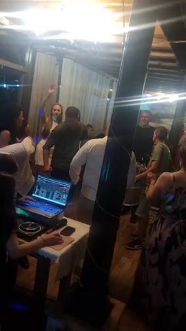 The video shows a DJ playing music at an indoor party. A group of people is gathered around the DJ, who is using a laptop and mixer on a table covered with a white cloth. Some people are dancing, while others are standing around talking. The room has a wooden floor and ceiling beams, with white curtains lining one wall. A large black pole is situated in the middle of the room. The lighting is bright and appears artificial.