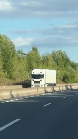 A white Scania semi-trailer truck drives on a highway. The truck is white with a long, light-colored trailer. The truck has a blue and white Scania logo on the front. The truck is driving on a highway with a concrete barrier to the right. Trees and foliage are visible behind the truck and barrier. The sky is partly cloudy.