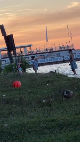 The video shows a grassy embankment next to a body of water, with a boat docked in the background. A large, white boat with a blue stripe is visible, and it appears to have people on deck. The word "PATENTU" is visible on the side of the boat, suggesting it is a charter or tour boat. The sky is a warm orange and pink, indicating either sunrise or sunset.

On the grassy area, there are a few people. To the left, a young person in swimwear is crouched down. In the center, an elderly person in a light blue shirt and shorts stands near the water's edge. To the right, a young person in a blue dress stands in shallow water.

In the foreground, a bright orange basketball lies on the grass. A duck is foraging in the grass, and another duck is visible in the water. The overall scene suggests a relaxed, outdoor setting during a picturesque time of day.