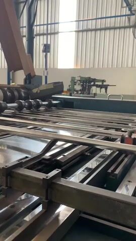 A conveyor belt system in a factory setting is shown. The conveyor belt consists of multiple parallel metal rollers and is transporting what appears to be flat, metallic sheets or large pieces of cardboard. To the left of the conveyor, a brown cardboard box is partially visible, leaning against some machinery. In the background, there is industrial equipment, including what looks like a printing press or a similar manufacturing machine with multiple arms and rollers. The environment is a well-lit factory with corrugated metal walls and ceiling. The video captures the smooth movement of the material on the conveyor belt.