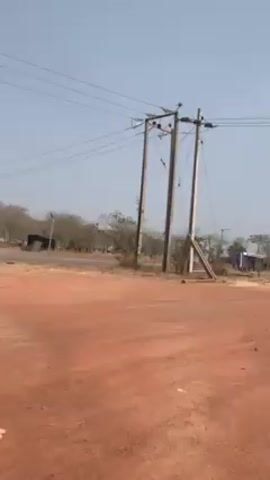 The video shows a dirt road with red soil. There are power lines and poles on the right. In the background, there's a communication tower, trees, and what seems to be a small building. The sky is clear and a light blue. There are no people visible in the video. The camera slowly pans from right to left.