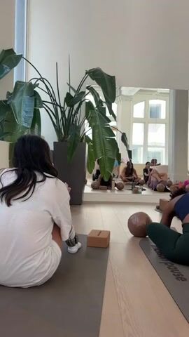 The video shows a group of women in a bright, airy studio, likely for a Pilates or yoga class. The room has a polished wooden floor, large mirrors reflecting the space, and high ceilings with decorative molding. Large potted plants add a touch of greenery. Various pieces of Pilates equipment, with pink accents, are visible in the background, suggesting a specialized fitness studio.

Several women are on yoga mats, engaged in exercises. In the foreground, a woman with dark curly hair is seen from behind, wearing a dark blue top and green leggings, sitting with her legs crossed on her mat. Another woman in a white top and dark shorts is also on a mat, facing away from the camera. In the middle ground, two women are lying on their backs on mats, performing a stretching exercise where they hold their knees to their chests. One wears a vibrant pink and white tie-dye top, and the other a bright pink sports bra. Small foam blocks and round foam rollers are scattered on the mats, common accessories for these types of workouts. The lighting is bright and natural, suggesting it could be daytime. The overall atmosphere appears calm and focused, consistent with a fitness class setting. The text "Mademoiselle Pause" is visible on the yoga mats. The location is identified as Montreal, Canada.