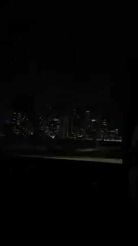 A dark video shot from inside a car shows a city skyline at night. The illuminated buildings of the cityscape are visible in the distance through the car window, contrasted by the darkness surrounding the vehicle. Some plants can be seen outside. The car appears to be moving, as indicated by the blurred movement of the city lights and roadside objects. A voice speaks in the background.