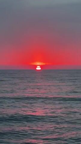 A close-up, vertical shot captures a vibrant red sun setting over a calm ocean. The sun, a brilliant orb of crimson, is partially submerged below the horizon line, casting a fiery glow across the sky and reflecting a shimmering path of light onto the water. The ocean's surface is a mosaic of gentle waves, their crests tinged with pink and purple hues from the sunset's illumination. The sky above transitions from the intense red of the sunset to a muted, hazy gray at the top of the frame. The scene is devoid of any human presence or man-made objects, offering a serene and unobstructed view of nature's spectacle.