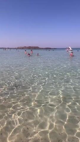 This is a vertical, first-person perspective video of a crowded beach on a clear, sunny day. The camera is positioned in the shallow, crystal-clear turquoise water, with sunlight glinting off the sandy bottom. In the foreground, small fish are visible swimming. Several people are scattered throughout the water, wading and swimming. In the middle ground, a group of people are gathered further out, and a white inflatable water slide structure is visible. In the background, more people are on a distant, rocky island or peninsula. Several boats and other watercraft are present on the horizon, including a white boat with people on it and a yellow inflatable watercraft. The sky is a deep, cloudless blue.