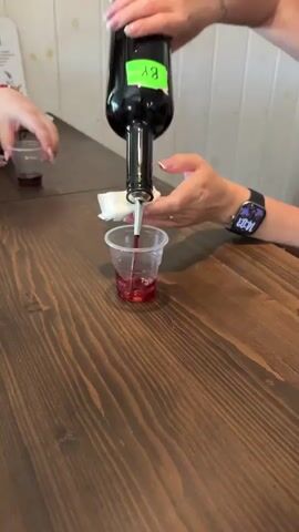 The video, likely filmed indoors in Rassvet, Russia, shows a person pouring a dark red liquid into a small clear plastic cup. The individual, wearing a black tank top and a smartwatch displaying "14:03," carefully pours from a dark green bottle fitted with a pour spout, which is wrapped in a white napkin. The liquid appears to be wine or a similar beverage. Another person's hands holding a cup are briefly visible in the background. The setting features a dark wooden planked table, a white wooden paneled wall, and a wooden chair. The audio includes a person speaking Russian, stating, "To us, girls, taste and aroma are pleasant, for men - presence," suggesting a discussion about the qualities of the drink being served.