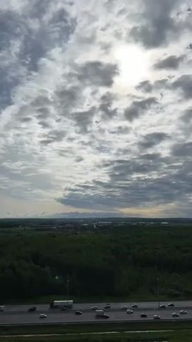The video shows an elevated view of a highway, a dense green forest, and a cityscape in the distance under a cloudy sky. The sun is obscured by the clouds but shines through, creating a bright spot. The highway has multiple lanes with cars and a truck moving along it. The forest stretches across the middle ground. The cityscape is faint on the horizon. The clouds are varied in shade, from light gray to darker storm clouds.