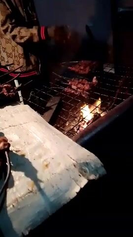 The video shows a man in a patterned jacket grilling meat over a fire. He pours liquid from a bottle onto the grilling meat. Skewers of meat are visible nearby. The grill sits on top of what appears to be a metal stand covered with white paper. A sign with the words "KARAOKE" and "LORIA KITCHEN" is visible in the background. The scene is dimly lit.