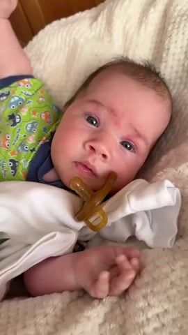 The video shows a close-up, eye-level shot of a newborn baby lying on a soft, textured blanket. The baby is wearing a white onesie with a green top underneath that has a print of cars and owls. A brown pacifier is in the baby's mouth. The baby's face is slightly red, and they appear to be looking at the camera with their eyes open. The background consists of a wooden surface, likely part of a crib or furniture. The lighting appears natural and soft. There is no discernible audio. The camera is held steady, with only slight movements as the baby's mouth moves around the pacifier. The baby's right hand is visible, with fingers slightly curled.