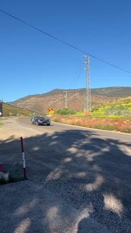 A black sports car drives on a paved road curving to the left in a rural setting under a clear blue sky. Mountains are visible in the background with vegetation and yellow flowers. Power lines run overhead. A road sign is visible on the curve. The camera is positioned on the side of the road, capturing the car as it moves past. The foreground includes shadows of trees or foliage.