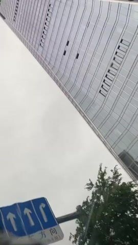 The video, shot from a moving vehicle, shows a modern glass skyscraper from a low angle as the car drives by. The sky is overcast and appears hazy. The building has a distinct curved facade. Reflecting in the glass are other buildings and the cloudy sky. On the lower portion of the skyscraper, there is a banner with Chinese characters and what appear to be floral graphics. Below the banner, text reads "办公热线 6666". A street sign with an arrow pointing upwards and a green pedestrian signal are visible in the foreground at the bottom of the frame. Trees can be seen to the right of the building. The car's blue bodywork is partially visible at the bottom. The footage is slightly shaky, consistent with being filmed from a moving vehicle.
